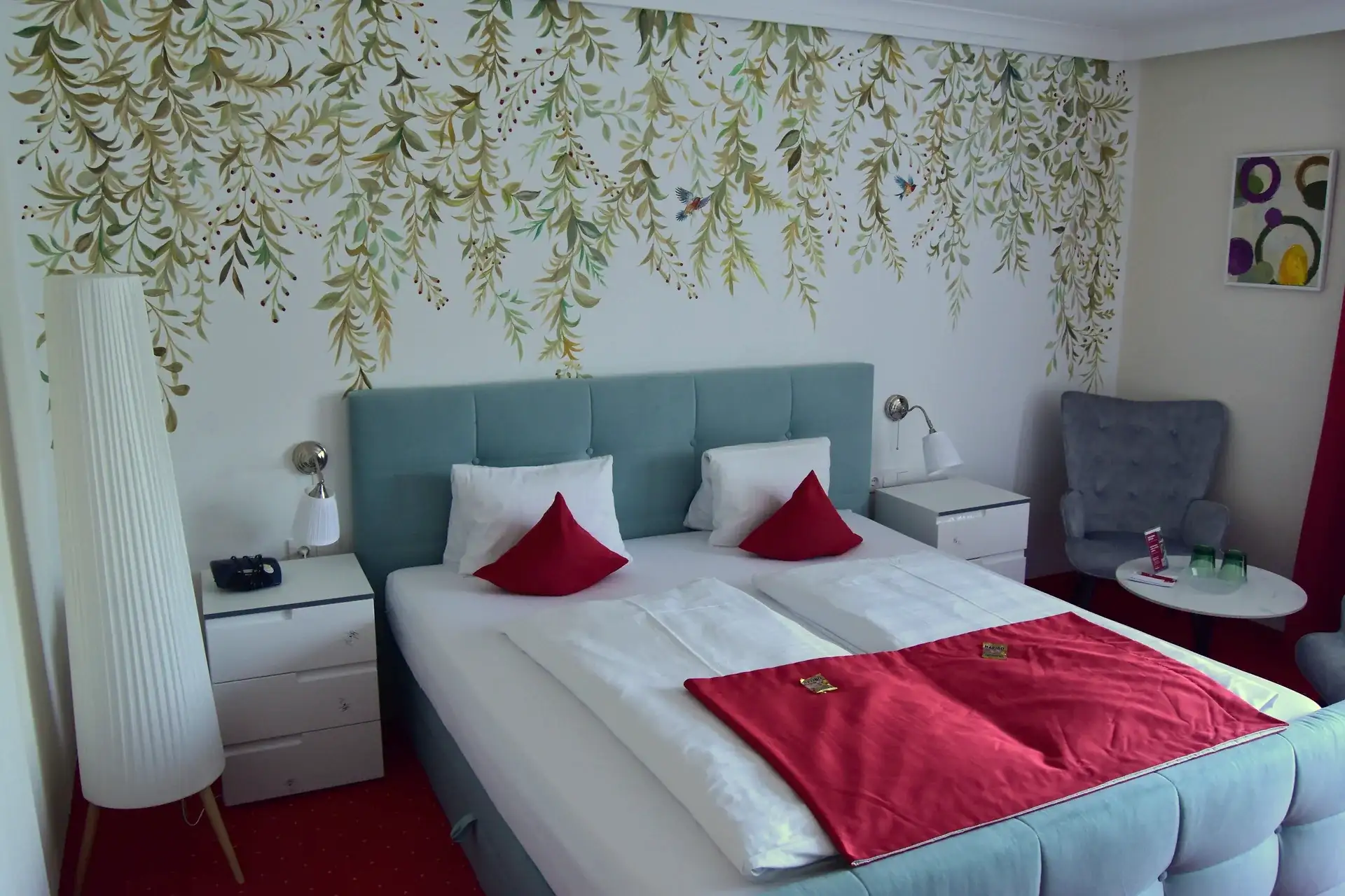 Doppelzimmer mit botanischer Wandgestaltung, gepolstertem Bett und roten Kissen